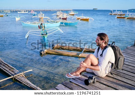 Photography and travel. Young man with rucksack holding camera sitting on wooden fishing pier with beautiful tropical sea view.