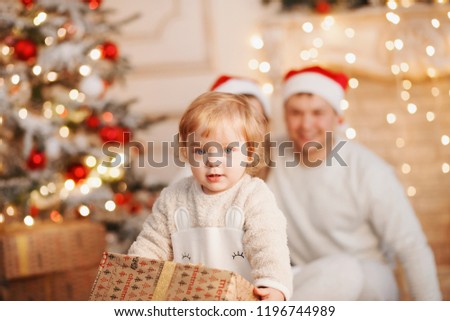 child opens the gift for Christmas. Young couple watch their daughter open her Christmas gift. Decorated Christmas interior at the background. Happy family picture