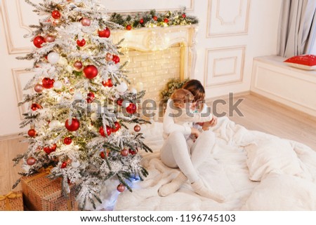 Young mother and her baby have fun sitting on a floor near white fireplace and Christmas tree with red Christmas bauble in anticipation of new year at home. First Christmas for baby.
