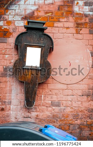 Dead mirror in a music instrument shaped frame hangs on red painted brick wall. Vertical. Dumpster in foreground