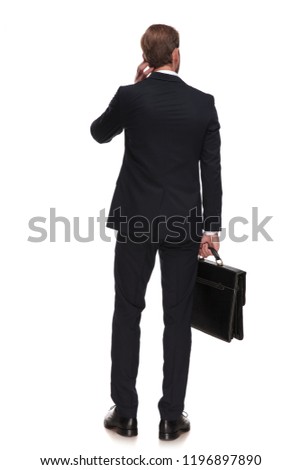 behind of pensive businessman holding suitcase and looking to side while standing on white background and touching his head, full body picture