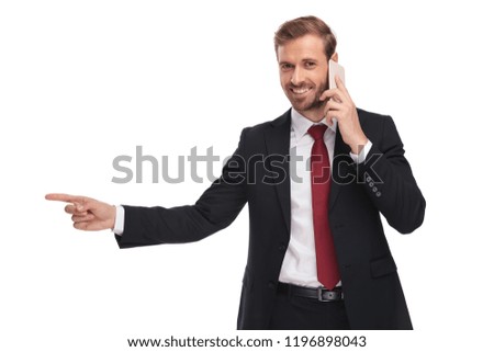 portrait of young businessman wearing a suit and a red tie talking on the phone and pointing to side while standing on white background