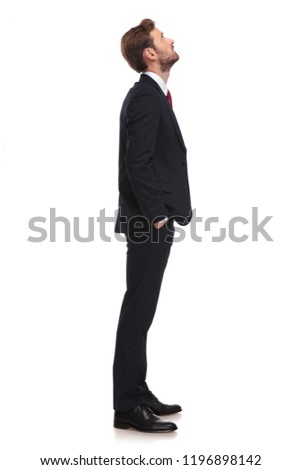young businessman waiting in line looks up at something while standing on white background with hands in pockets, full length picture