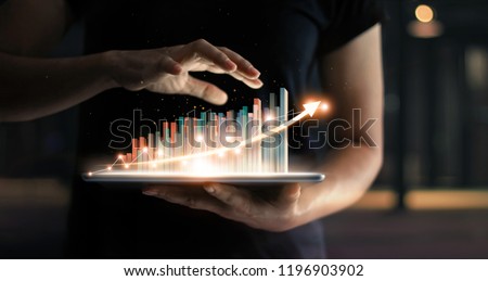businessman holding tablet and showing a growing virtual hologram of statistics, graph and chart with arrow up on dark background. Stock market. Business growth, planing and strategy concept.  Royalty-Free Stock Photo #1196903902