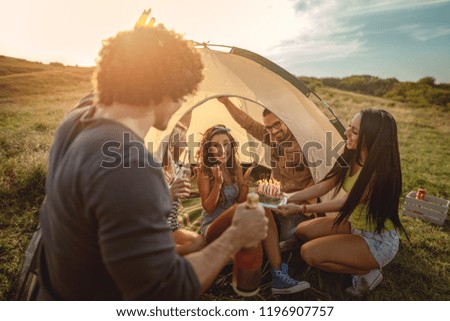 Young people have a good time in camp in nature. They're celebrating a birthday, laughing and greeting to their friend with birthday cake, happy to be together.