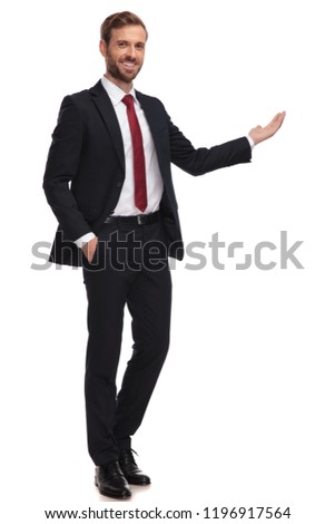smiling businessman stands on white background with hand in pocket and presents to side, full length picture
