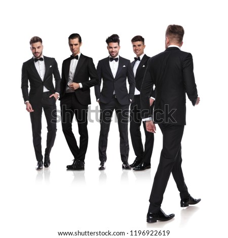 five men dressed in tuxedoes with leader walking back to side and looking at them, on white background, full length picture