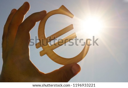 man's hand hold the Euro icon silhouette against sunny orange golden and yellow sky. sun rays. euro sign, symbol of money, idea of Euro Union, sun rays beam.