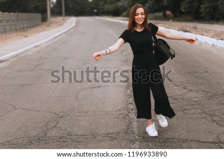 Full-length portrait of fashion stylish girl in the city. She walks on the street and turn around on camera. Dressed in black t-shirt, pants and white sneakers. Truly emotions. Place for text.