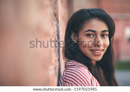 Dark haired woman. Pretty long haired young woman standing against the brick wall and smiling friendly while looking at you
