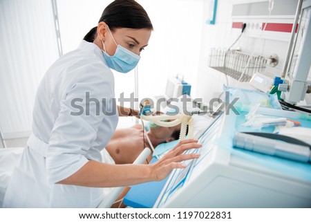 Waist up portrait of female medical worker in protective mask touching monitor of mechanical ventilator. Man lying in hospital bed on blurred background Royalty-Free Stock Photo #1197022831