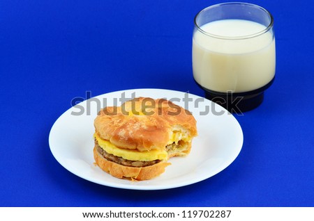Sausage and egg croissant with cheese and glass of milk on blue background.  Bite taken from sandwich.
