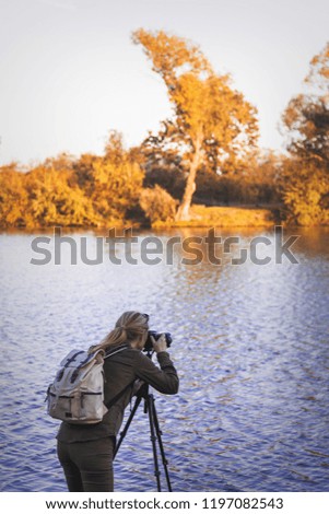 Woman is taking picture of landscape at autumn. Hiker with camera on tripod standing next to lake. 