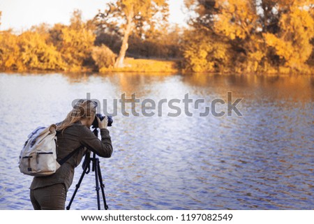 Woman is taking picture of landscape at autumn. Hiker with camera on tripod standing next to lake.