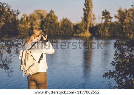 Woman with backpack is photographing a landscape near to lake, toned picture. Hiking woman in nature. 