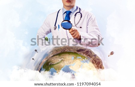 Close up of young doctor in white medical uniform looking through loupe and studying Earth globe, with cloudy skyscape on background. Medical industry concept. Elements of this image furnished by NASA