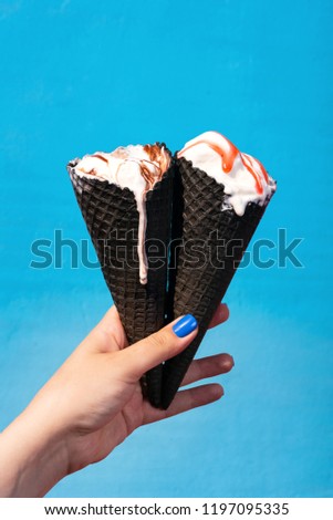 Hand holding two black ice cream cones with ice cream slightly melting in the sun on light blue background