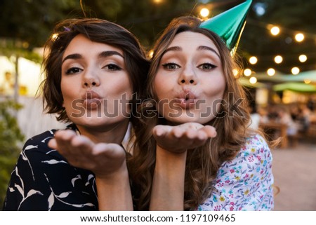 Image of happy young women friends outdoors in park having fun looking camera take a selfie blowing kisses.