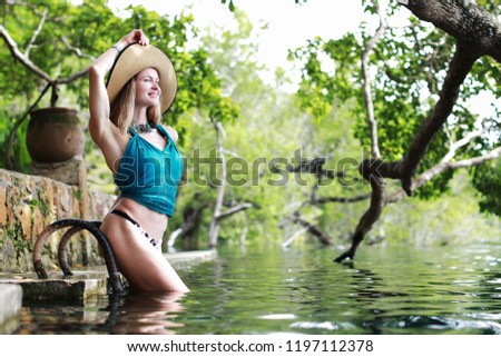 beautiful blonde woman in summer hat and aquamarine swimsuit posing near nature laguna in Mexico in summertime at green forest background 