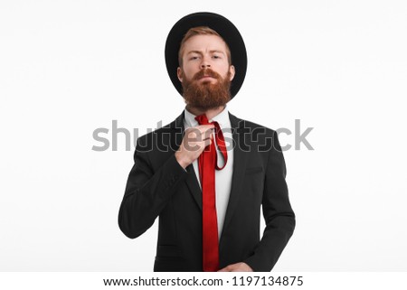Style, men's wear and fashion concept. Picture of fashionable Caucasian male with thick ginger beard getting dressed for some official event, wearing black hat and suit, knotting red elegant tie