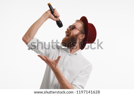 People, music, fun, show and entertainment concept. Studio shot of emotional handsome stylish red haired pop artist with thick beard singing in microphone wearing red round hat and trendy shades