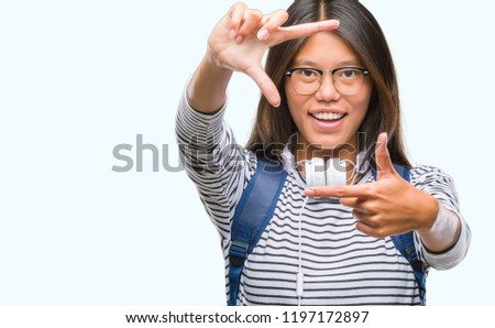 Young asian student woman wearing headphones and backpack over isolated background smiling making frame with hands and fingers with happy face. Creativity and photography concept.