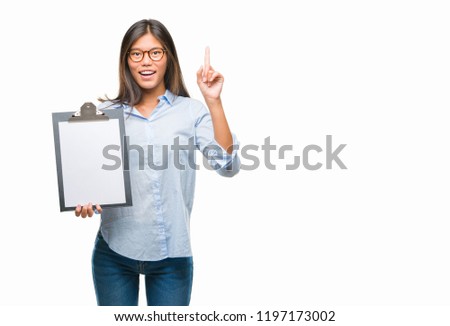 Young asian business woman over isolated background holding clipboard surprised with an idea or question pointing finger with happy face, number one