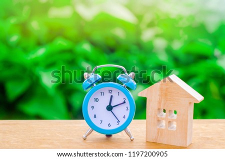 Wooden house and blue alarm clock on a green background. The concept of rent housing monthly and hourly. Temporary affordable accommodation, hotels and hostels. A loan to buy a house.