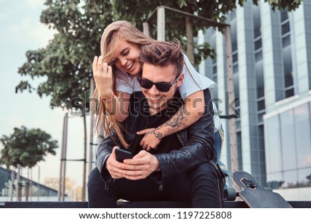 Attractive trendy dressed young couple sitting together on bench near skyscraper - pretty girl embraces her boyfriend.