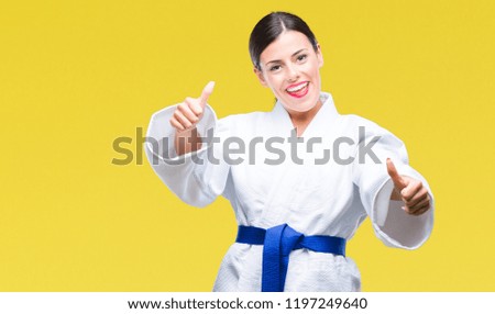 Young beautiful woman wearing karate kimono uniform over isolated background approving doing positive gesture with hand, thumbs up smiling and happy for success. Looking at the camera, winner gesture.