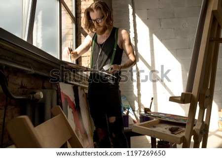 Portrait of handsome male artist painting picture standing by window art studio lit by sunlight, copy space