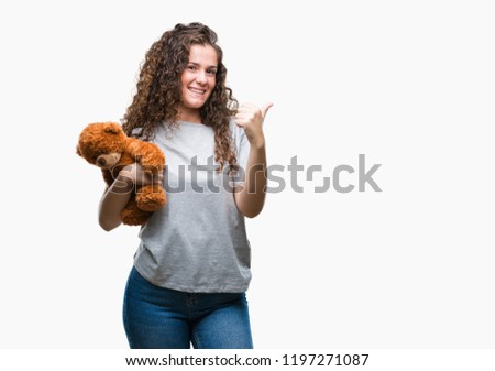 Young brunette girl holding teddy bear over isolated background happy with big smile doing ok sign, thumb up with fingers, excellent sign