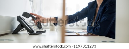 View through an office partition of a businesswoman or secretary picking or hanging up telephone handset. Royalty-Free Stock Photo #1197311293