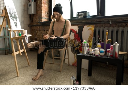 Full length portrait of handsome contemporary artist drawing sketches while sitting by easel in loft like art studio, copy space