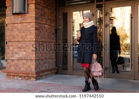 Young female student in warm autumn clothes coming out of the house with a backpack with books and a cup of coffee. Autumn trend, urban stydent style.
