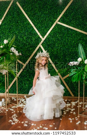 Cute little girl with blond curly hair in a white wedding dress and a wreath of flowers in floral decorations made from tropical leaves, white roses and orchids
