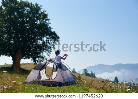 Young pretty woman standing on steep grassy hill at small tourist tent and big tree and taking picture of beautiful foggy mountains covered with forest under clear blue sky on bright summer morning.