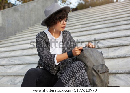 Young attractive female, wears cardigan, shirt, hat and glasses, with curly dark hair, open her backpack and searching something inside.
