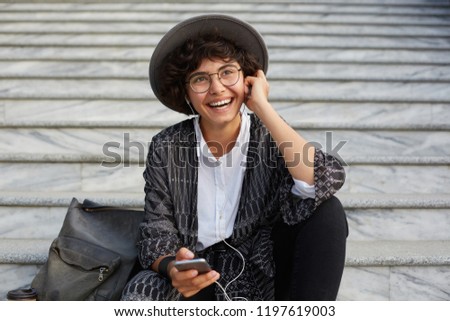 outdoor shot of young attractive woman wears white shirt cardigan and hat smiles and listen music via headphones. businesswoman sittin on stairs and feels happy