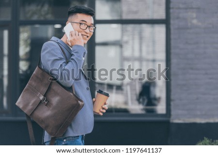 handsome asian man with leather backpack and coffee to go talking on smartphone 