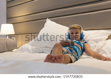 Smiling kid with headphones watching cartoons on smart phone while lying down on bed. 