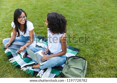Two smiling young girls students sitting on a grass at the campus, studying with laptop computers