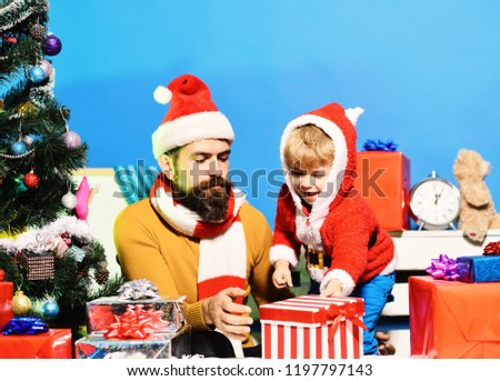 Santa and little assistant among gift boxes near Christmas tree. Family holidays concept. Man with beard and curious face plays with son. Christmas family opens presents on blue background.