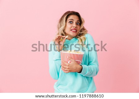 Image of displeased european woman 20s holding bucket with popcorn and looking at camera isolated over pink background