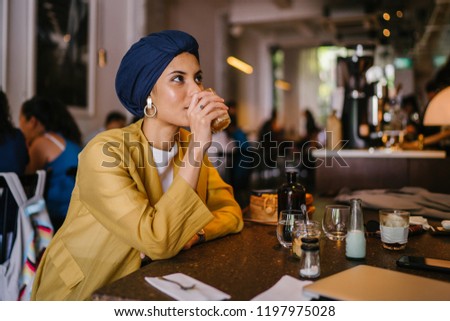 Portrait of a young and attractive Malay Muslim woman enjoying a meal and a cup of coffee in a cafe. She is wearing a turban (hijab, head scarf) and is tall, elegant and fashionably dressed. Royalty-Free Stock Photo #1197975028