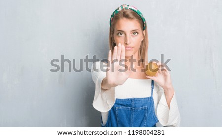 Beautiful young woman over grunge grey wall holding fresh kiwi with open hand doing stop sign with serious and confident expression, defense gesture