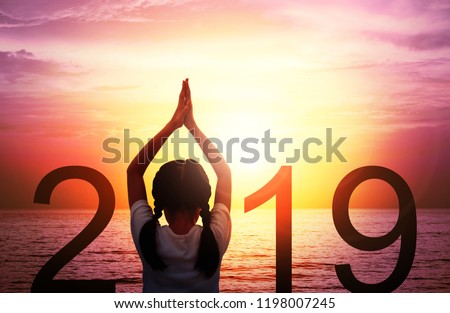 Happy new year card 2019. Silhouette of girl doing Yoga vrikshasana tree pose on tropical beach with fantastic sunset sky background. Kid standing as part of the Number 2019 sign and watching sunrise.