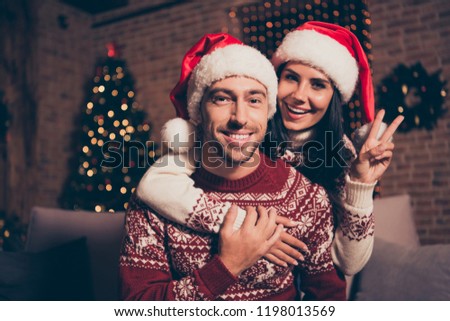Noel christmastime concept. Two beautiful, sweet, charming people in ornament sweater sit in comfort, cozy sofa or couch in living room with sparkles garland decorations on pine tree make v-sign