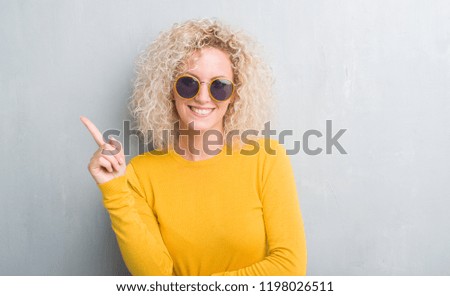 Young blonde woman with curly hair over grunge grey background with a big smile on face, pointing with hand and finger to the side looking at the camera.