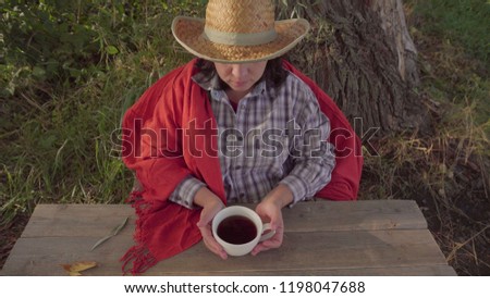 A village woman drinks tea. Cowboy straw hat. On the background of a large tree. The view from the top. Green grass and leaves. Country life. Serene atmosphere.